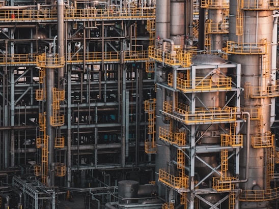 An intricate network of industrial metal structures and machinery with several tall cylindrical towers interconnected by yellow safety railings. The complex design and metallic components indicate a large-scale industrial facility, possibly a refinery or power plant.
