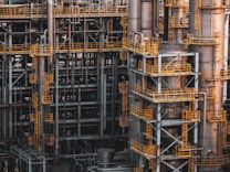 An intricate network of industrial metal structures and machinery with several tall cylindrical towers interconnected by yellow safety railings. The complex design and metallic components indicate a large-scale industrial facility, possibly a refinery or power plant.