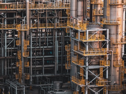 An intricate network of industrial metal structures and machinery with several tall cylindrical towers interconnected by yellow safety railings. The complex design and metallic components indicate a large-scale industrial facility, possibly a refinery or power plant.