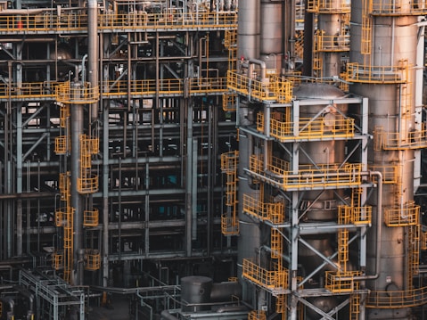An intricate network of industrial metal structures and machinery with several tall cylindrical towers interconnected by yellow safety railings. The complex design and metallic components indicate a large-scale industrial facility, possibly a refinery or power plant.