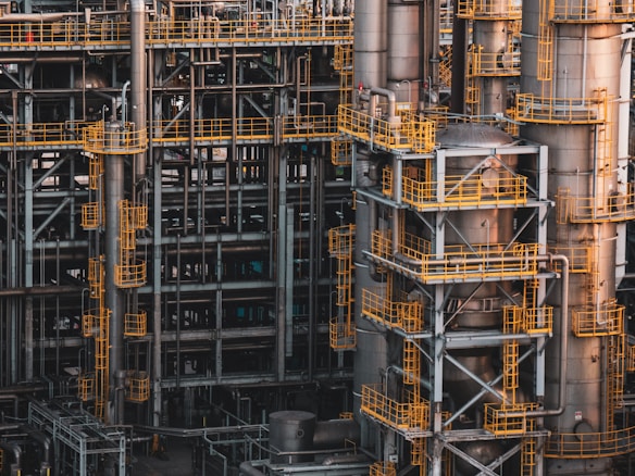 An intricate network of industrial metal structures and machinery with several tall cylindrical towers interconnected by yellow safety railings. The complex design and metallic components indicate a large-scale industrial facility, possibly a refinery or power plant.