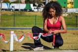 A young woman with curly hair, wearing a red top and sunglasses, is kneeling on grass while tying colorful streamers onto a pink cord. The setting is an outdoor area with some trees and trucks visible in the background, suggesting a park or open field.
