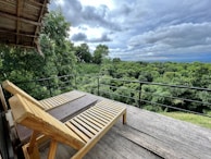 A wooden deck featuring a reclining lounge chair overlooks a lush, green forest. The sky is overcast with layers of thick clouds, creating a serene and tranquil atmosphere. The deck is bordered by a simple black railing, providing a seamless view of the expansive treetops and dense foliage.