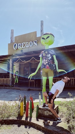 A person is playfully posing with a large green alien statue wearing a patterned shirt in front of a rustic building labeled 'OREGON'. The scene includes colorful, oversized crayon-like objects in the foreground and has a bright, sunny ambiance.
