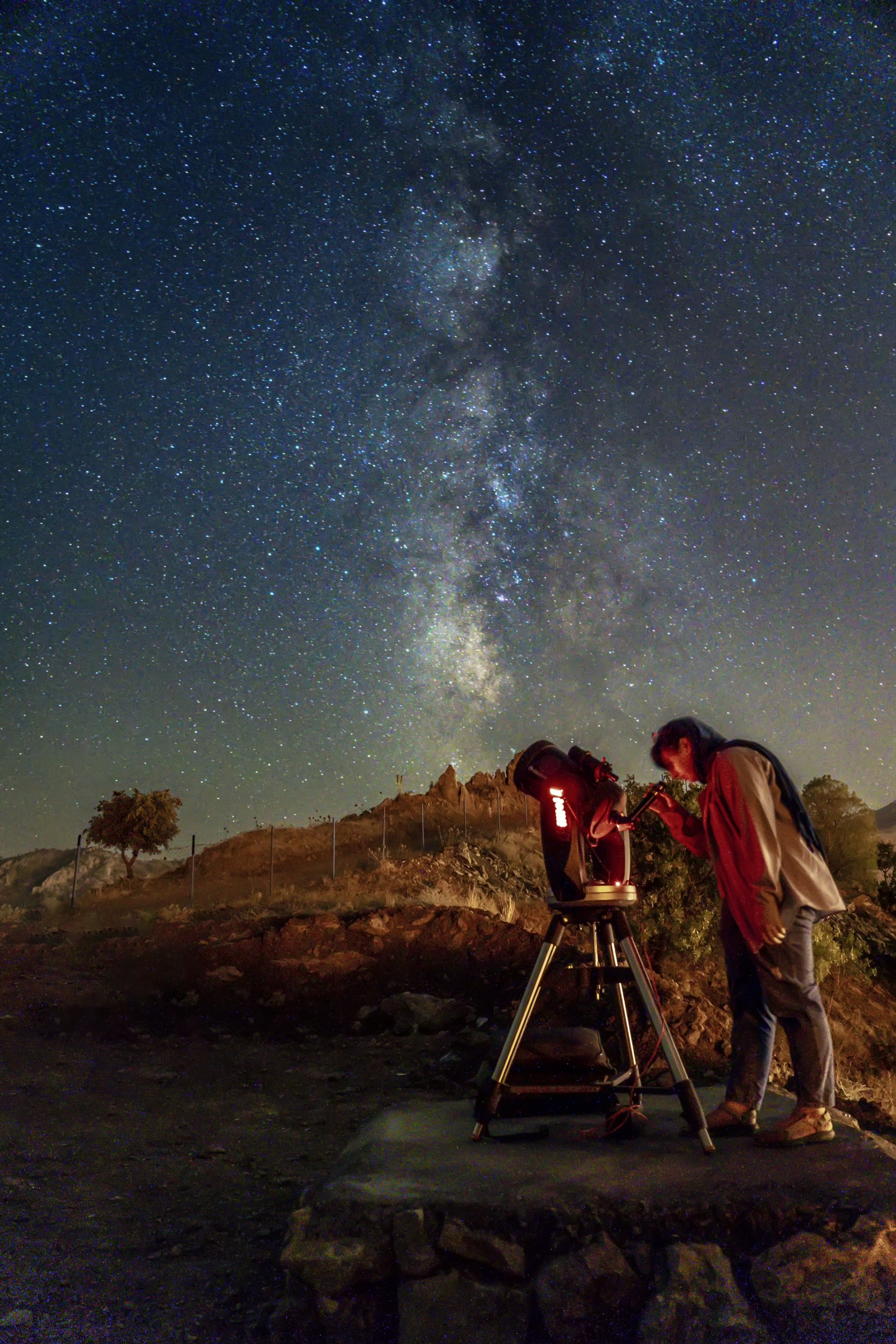 Person looking through telescope at night in open desert, Milky Way visible, dark moody atmosphere, low ambient light