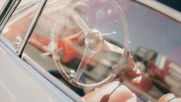 A classic car interior with a large, round metal steering wheel and shiny dashboard. The upholstery is a rich red color, and the sunlight creates a soft glow on the surfaces.