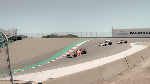 Three vintage race cars are navigating a curved section of a racetrack surrounded by fencing and asphalt barriers. The scene is set during daytime with clear, bright lighting, and there is a sign in the background indicating a motorsport event.