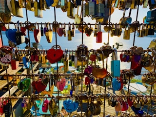A collection of various colorful locks attached to metal railings, with a background view of the sea and sunlight reflecting on the water. Some locks are heart-shaped, while others come in different shapes and sizes.