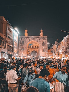 A crowded marketplace at night with a historic archway or gate in the background. The market is bustling with people, including some street vendors and shoppers. There are also illuminated buildings and streetlights adding to the vibrant atmosphere.