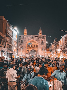 A crowded marketplace at night with a historic archway or gate in the background. The market is bustling with people, including some street vendors and shoppers. There are also illuminated buildings and streetlights adding to the vibrant atmosphere.