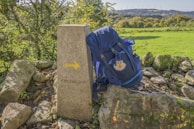 A backpack rests against a stone marker with a yellow arrow and the inscription 'Km. 100,000'. The scene is set in a rural area with lush green grass and a backdrop of hills and trees, suggesting a hiking or pilgrimage route.