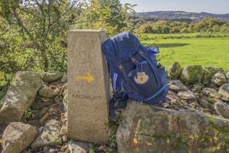 A backpack rests against a stone marker with a yellow arrow and the inscription 'Km. 100,000'. The scene is set in a rural area with lush green grass and a backdrop of hills and trees, suggesting a hiking or pilgrimage route.
