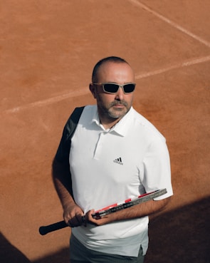 A man wearing sunglasses is holding a tennis racket on a clay tennis court. He is dressed in a white polo shirt and appears serious and focused.