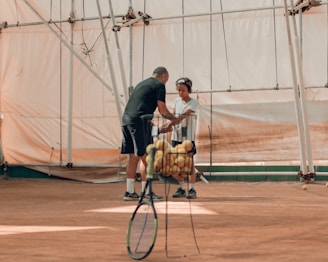 A tennis coach is giving instructions to a young player on an indoor clay court. There is a basket of tennis balls and a tennis racket in the foreground. The background shows the interior of the court structure with metal scaffolding and cream-colored fabric walls.