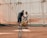 A tennis coach is giving instructions to a young player on an indoor clay court. There is a basket of tennis balls and a tennis racket in the foreground. The background shows the interior of the court structure with metal scaffolding and cream-colored fabric walls.
