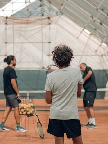 Three people are playing tennis indoors on a clay court. One person is holding a tennis racket, while another stands near a basket filled with tennis balls. The players are casually dressed in sports attire, and the surrounding structure features a high, arched ceiling with a mix of transparent and opaque panels.