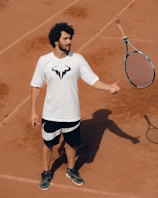 A person stands on a clay tennis court, wearing a white t-shirt with a logo and black shorts. A tennis racquet is being tossed or caught in the air, casting a shadow on the ground.