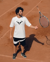 A person stands on a clay tennis court, wearing a white t-shirt with a logo and black shorts. A tennis racquet is being tossed or caught in the air, casting a shadow on the ground.