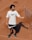 A person stands on a clay tennis court, wearing a white t-shirt with a logo and black shorts. A tennis racquet is being tossed or caught in the air, casting a shadow on the ground.