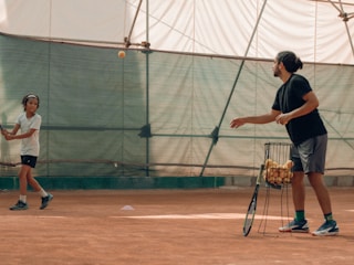 A young tennis player in sports attire is at a tennis court with their coach. The coach is holding a basket full of tennis balls and tossing one towards the player. They are both indoors, and the playing surface appears to be clay. The environment is well-lit, and the setting seems focused on training.