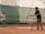 A young tennis player in sports attire is at a tennis court with their coach. The coach is holding a basket full of tennis balls and tossing one towards the player. They are both indoors, and the playing surface appears to be clay. The environment is well-lit, and the setting seems focused on training.