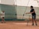 A young tennis player in sports attire is at a tennis court with their coach. The coach is holding a basket full of tennis balls and tossing one towards the player. They are both indoors, and the playing surface appears to be clay. The environment is well-lit, and the setting seems focused on training.