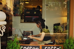 A young woman is working on a laptop inside a cozy cafe. She appears focused, sitting at a small wooden table with empty glasses and a plant nearby. The interior is warmly lit with a mix of natural light from the window, through which the reflection of outdoor greenery and buildings can be seen.