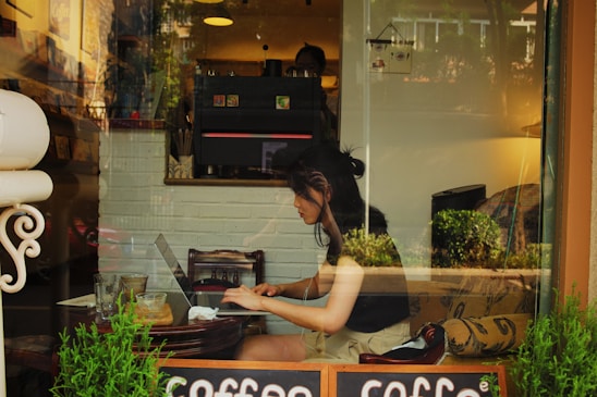 A young woman is working on a laptop inside a cozy cafe. She appears focused, sitting at a small wooden table with empty glasses and a plant nearby. The interior is warmly lit with a mix of natural light from the window, through which the reflection of outdoor greenery and buildings can be seen.