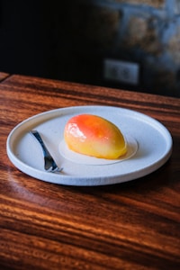 A pastry shaped like a mango sits elegantly on a white, round plate with a small fork to the side. The pastry has a glossy finish with hues of yellow and orange, resembling a ripe mango. The plate rests on a wooden tabletop, creating a rustic yet sophisticated presentation.