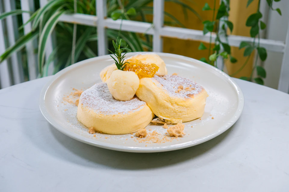The image shows a white plate with three golden brown pancakes on it, each topped with a dollop of cream and a sprig of rosemary. The plate is placed on a table covered with a white tablecloth, which contrasts with the warm tones of the pancakes. In the background, there are green plants adding a touch of nature to the scene.