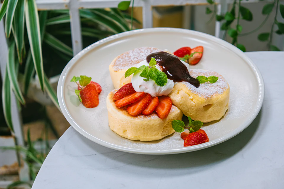 The image shows a white plate with three golden brown pancakes topped with fresh strawberries and whipped cream, resting on a white tablecloth against a blurred background.