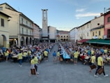 A large outdoor gathering in a town square with people seated at long tables. Many attendees are wearing yellow shirts, suggesting an organized event or festival. Strings of blue and white streamers hang overhead. The square is lined with colorful buildings and an archway leads to a tower. There are people serving food and drinks, and the atmosphere is bustling with activity.