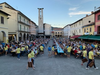 A large outdoor gathering in a town square with people seated at long tables. Many attendees are wearing yellow shirts, suggesting an organized event or festival. Strings of blue and white streamers hang overhead. The square is lined with colorful buildings and an archway leads to a tower. There are people serving food and drinks, and the atmosphere is bustling with activity.