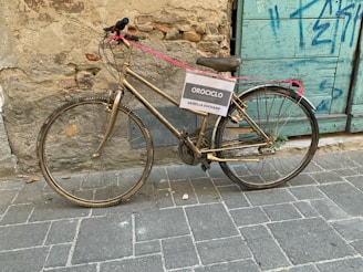 A golden bicycle is securely locked with a pink cable against an aged brick wall with a rustic blue wooden door. The bicycle has a sign attached to it reading 'OROCICLO - GRISELLA DUCHAMP.' The surrounding area features graffiti and a paved stone walkway with some fallen leaves.