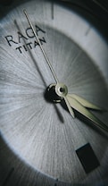The close-up shot captures the intricate details of a watch face featuring the words 'RAGA TITAN'. The watch hands are sharply focused against a textured, metallic background, emphasizing precision and craftsmanship.