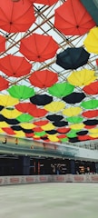 A vibrant display of red, yellow, green, and black umbrellas attached to the ceiling in an indoor space. Below the umbrellas, a skating rink can be seen partially, with a sign advertising umbrellas along the perimeter. The ceiling beams crisscross in a lattice formation, creating an artistic canopy of varied colors.