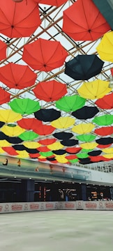 A vibrant display of red, yellow, green, and black umbrellas attached to the ceiling in an indoor space. Below the umbrellas, a skating rink can be seen partially, with a sign advertising umbrellas along the perimeter. The ceiling beams crisscross in a lattice formation, creating an artistic canopy of varied colors.