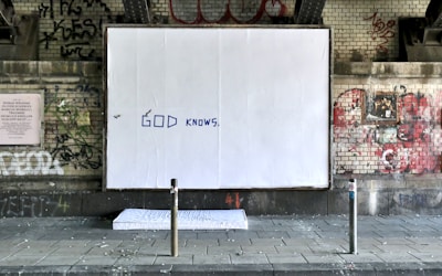 A large white billboard with the words 'GOD KNOWS' written in blue is located under a graffiti-covered brick wall. The setting appears to be urban, possibly beneath a bridge or overpass. A mattress lies on the ground in front of the billboard. The environment looks grungy with various tags and street art surrounding the main focus.