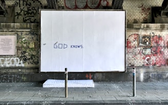 A large white billboard with the words 'GOD KNOWS' written in blue is located under a graffiti-covered brick wall. The setting appears to be urban, possibly beneath a bridge or overpass. A mattress lies on the ground in front of the billboard. The environment looks grungy with various tags and street art surrounding the main focus.