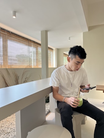 A person sits indoors at a modern, minimalistic setting with large windows and natural light. They are casually dressed in a white t-shirt and black pants, holding and looking at a smartphone. In their other hand, they hold a glass containing a green-colored beverage with ice and a straw.