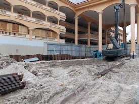 A construction site with a large building featuring multiple arches and columns in the background. The foreground is dominated by piles of dirt and construction equipment, including an excavator. There are metal beams and supporting structures scattered around, and a person wearing a safety helmet is standing nearby.