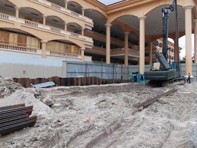 A construction site with a large building featuring multiple arches and columns in the background. The foreground is dominated by piles of dirt and construction equipment, including an excavator. There are metal beams and supporting structures scattered around, and a person wearing a safety helmet is standing nearby.