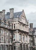 An ornate, historic stone building features intricate architectural details such as high-pitched roofs, dormer windows, and carved embellishments. Large windows with small panes are set in symmetrical patterns, and the overall aesthetic is Gothic Revival, suggesting an education or institutional purpose.