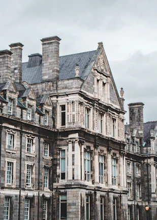 An ornate, historic stone building features intricate architectural details such as high-pitched roofs, dormer windows, and carved embellishments. Large windows with small panes are set in symmetrical patterns, and the overall aesthetic is Gothic Revival, suggesting an education or institutional purpose.