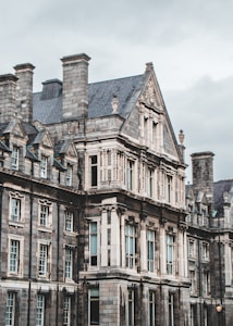 An ornate, historic stone building features intricate architectural details such as high-pitched roofs, dormer windows, and carved embellishments. Large windows with small panes are set in symmetrical patterns, and the overall aesthetic is Gothic Revival, suggesting an education or institutional purpose.
