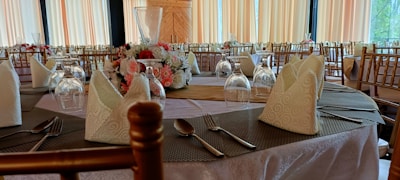 A beautifully set banquet table is adorned with elegant folded napkins, neatly arranged glassware, and a floral centerpiece featuring roses and other flowers. The decor includes wooden chairs and light-colored tablecloths, creating a sophisticated and inviting dining atmosphere.