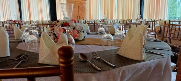 A beautifully set banquet table is adorned with elegant folded napkins, neatly arranged glassware, and a floral centerpiece featuring roses and other flowers. The decor includes wooden chairs and light-colored tablecloths, creating a sophisticated and inviting dining atmosphere.