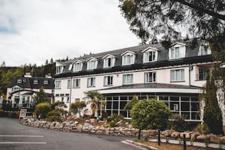 A large building with multiple windows and a dark roof, surrounded by lush greenery and a well-maintained garden. There are several smaller plants and rocks arranged neatly in front of the building, which appears to be a hotel or a similar establishment. The road in front and a parking space are visible, with a sign indicating that the place is open. Tall trees are in the background, and the sky is overcast, suggesting a calm and serene atmosphere.