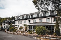 A large building with multiple windows and a dark roof, surrounded by lush greenery and a well-maintained garden. There are several smaller plants and rocks arranged neatly in front of the building, which appears to be a hotel or a similar establishment. The road in front and a parking space are visible, with a sign indicating that the place is open. Tall trees are in the background, and the sky is overcast, suggesting a calm and serene atmosphere.