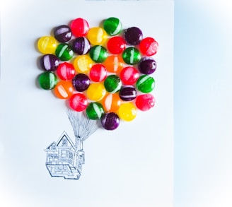 A playful arrangement of colorful candies placed strategically on a piece of paper, resembling balloons lifting a hand-drawn sketch of a house. The candies are vibrant and round, creating a whimsical and imaginative scene.